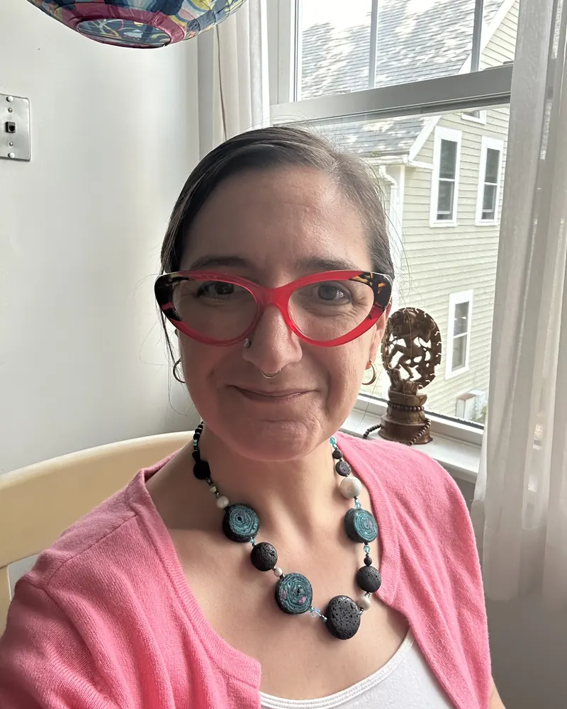 Kara Katz by a sunny office window in red glasses and a beaded necklace